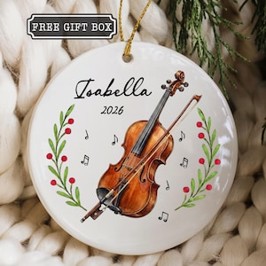 May include: A round white ceramic ornament featuring a watercolor illustration of a brown violin with a bow, surrounded by green leafy branches with red berries and black musical notes. The text Isabella and 2026 are printed in black ink.