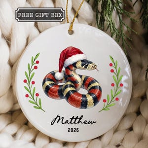 May include: A round white ceramic ornament featuring a coiled snake with red, black, and yellow bands wearing a red and white Santa hat. The design includes green holly branches with red berries, the name Matthew, and the year 2026 in black text.