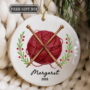 May include: A round white ceramic ornament featuring a red yarn ball with two crossed wooden knitting needles. The design is framed by green leafy sprigs with red berries. The text reads Margaret and 2026, with a label stating FREE GIFT BOX.