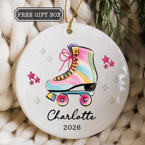 May include: A round white ceramic ornament featuring a colorful roller skate illustration with pink laces, surrounded by small pink stars and sparkles. The name Charlotte and the year 2026 are printed in black text below the roller skate graphic.