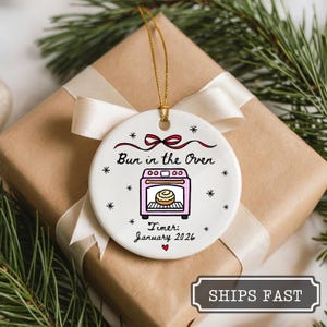 K&ouml;nnte beinhalten: Wei&szlig;er Keramik-Weihnachtsschmuck mit dem Text "Bun in the Oven" und einer rosa Ofenillustration. Der Schmuck enth&auml;lt den Text "Timer: January 2026" und ist mit Schneeflocken und einer roten Schleife verziert. Er h&auml;ngt an einer goldenen Schnur.