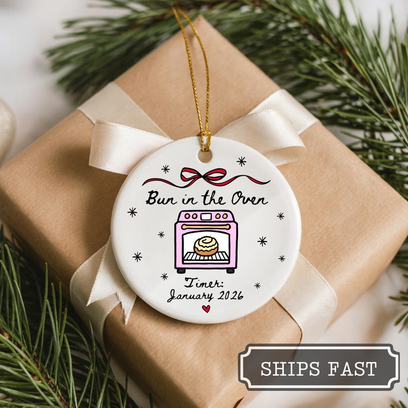 K&ouml;nnte beinhalten: Wei&szlig;er Keramik-Weihnachtsschmuck mit dem Text "Bun in the Oven" und einer rosa Ofenillustration. Der Schmuck enth&auml;lt den Text "Timer: January 2026" und ist mit Schneeflocken und einer roten Schleife verziert. Er h&auml;ngt an einer goldenen Schnur.