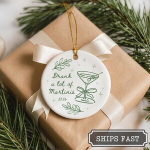 May include: A round white ceramic ornament featuring a green line drawing of a martini glass with olives, a bow, and leafy sprigs. The text reads Drank a lot of Martinis and 2026, accented by small star shapes. A gold string is attached for hanging.