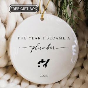 May include: A round white ceramic ornament featuring the black text THE YEAR I BECAME A plumber, a black faucet and wrench icon, and the year 2026. A gold string is attached to the top for hanging. A small rectangular label reads FREE GIFT BOX.