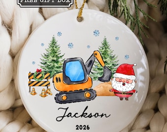 Pode incluir: Um ornamento redondo de cerâmica branca com um cordão dourado. O design inclui uma escavadora laranja, um Pai Natal, pinheiros verdes e cones de construção. O texto diz "FREE GIFT BOX", "Jackson" e "2026".