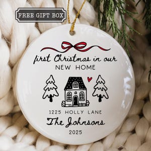 May include: A white ceramic Christmas ornament with a gold string. The ornament features the text "first Christmas in our NEW HOME" with a house and trees illustration. It also includes the address "1225 HOLLY LANE", the name "The Johnsons", and the year "2025".
