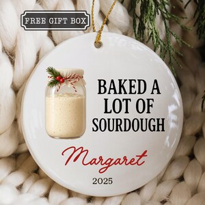 May include: White ceramic ornament with a jar of sourdough bread and the text "BAKED A LOT OF SOURDOUGH Margaret 2025". The ornament has a gold string for hanging and a small sprig of greenery.