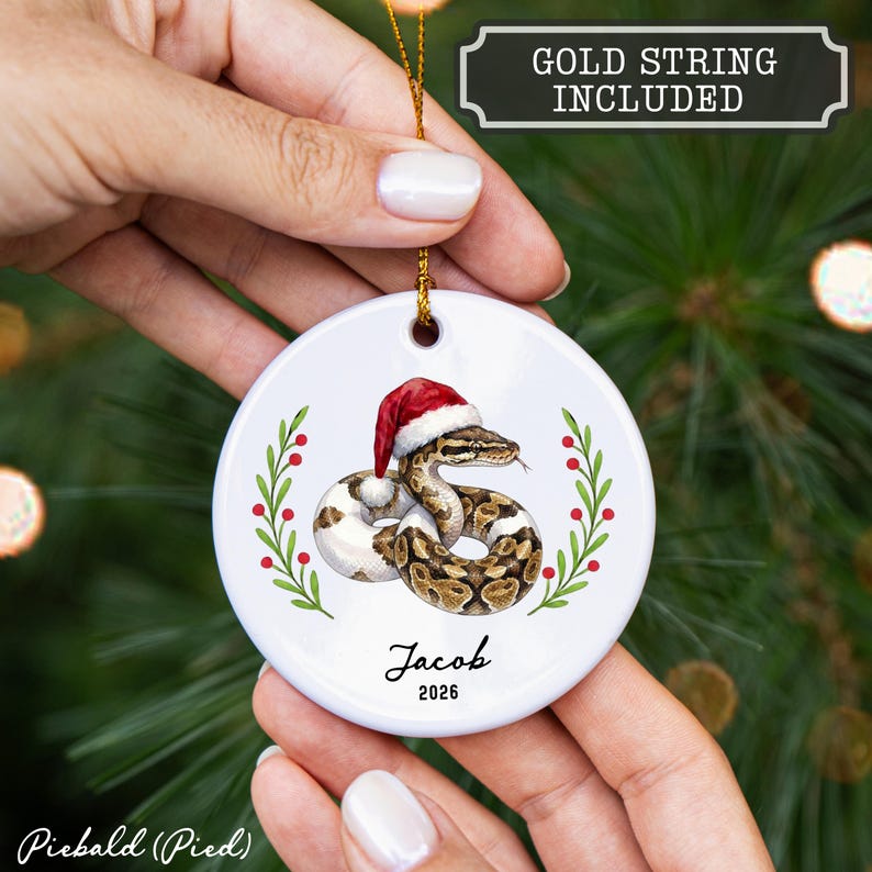 May include: A round white ceramic ornament featuring a piebald ball python wearing a red and white Santa hat, flanked by green holly branches with red berries. The text reads Jacob and 2026. A gold string is included for hanging the ornament.