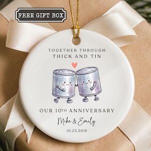 May include: A round white ceramic ornament featuring a gold hanging string. The design shows two illustrated tin cans holding hands with a small red heart above them. Text reads: TOGETHER THROUGH THICK AND TIN, OUR 10TH ANNIVERSARY, Mike & Emily, 10.23.2016.