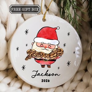 May include: A round white ceramic ornament featuring a cartoon Santa Claus holding a brown patterned snake. The design includes black star accents, the name Jackson, and the year 2026 in black text. A gold string is attached to the top for hanging.