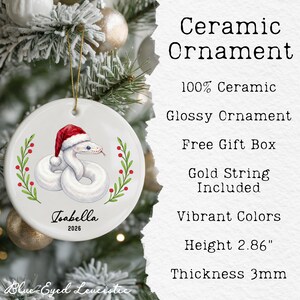 May include: A round white ceramic ornament featuring a graphic of a white snake wearing a red and white Santa hat, flanked by green holly branches with red berries. The text Isabella and 2026 are printed below. It includes a gold string and is 2.86 inches high.