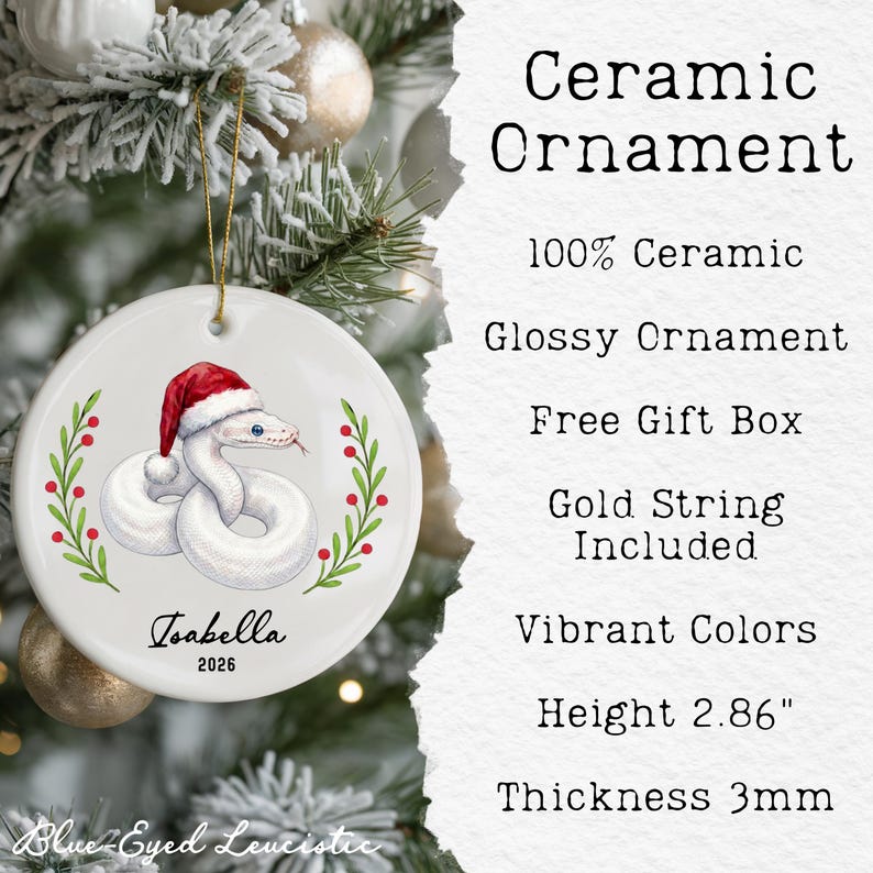 May include: A round white ceramic ornament featuring a graphic of a white snake wearing a red and white Santa hat, flanked by green holly branches with red berries. The text Isabella and 2026 are printed below. It includes a gold string and is 2.86 inches high.