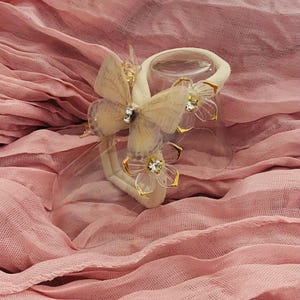 May include: A decorative hair accessory featuring a cream-colored band with a butterfly and flower embellishments. The butterfly has a tan body and wings, with a rhinestone center. The flowers have clear petals with gold trim and rhinestone centers. The accessory sits on a pink fabric.