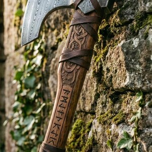Puede incluir: Hacha artesanal con una hoja de acero estampada y un mango de madera. El mango está intrincadamente tallado con diseños de nudos celtas y runas, envuelto en cuero marrón. El hacha está apoyada contra una pared de piedra cubierta de musgo.