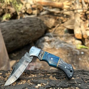 Puede incluir: Un cuchillo plegable parcialmente abierto con una hoja estampada y un mango de madera azul, gris y negra. El cuchillo tiene una virola plateada y está apoyado sobre un trozo de madera.