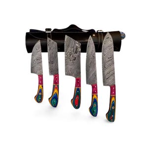 Puede incluir: Un juego de cinco cuchillos de cocina con hojas de acero estampadas y mangos de madera coloridos. Los cuchillos se exhiben en un estuche enrollable de cuero negro. Los mangos presentan una combinación de tonos rojos, amarillos, verdes y azules.