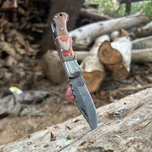 Puede incluir: Una navaja con una hoja estampada y un mango de madera. El mango presenta una combinación de acentos marrones, rojos y blancos. El cuchillo se muestra al aire libre, apoyado en un tronco.