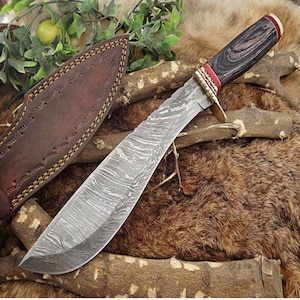 Puede incluir: Un cuchillo grande de acero de Damasco con mango de madera oscura, detalles rojos y guarda de latón. La hoja tiene un patrón distintivo. Una funda de cuero marrón es visible a la izquierda. El cuchillo descansa sobre un lecho de piel y madera.