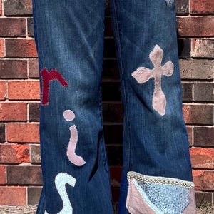 May include: Blue denim jeans with decorative patches and the word "rise" spelled out in various colors. The jeans feature cross-shaped patches and patchwork designs on the lower legs.