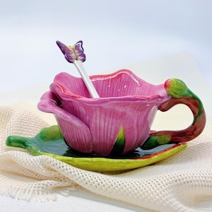 May include: A pink ceramic teacup and saucer set shaped like a flower. The teacup has a matching spoon with a butterfly on top. The handle is brown and green. The saucer is green and yellow. The set is on a white cloth.