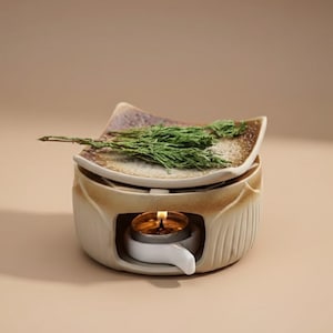 May include: A ceramic essential oil diffuser with a lit tea light candle. The diffuser is beige with a textured design and a small dish on top holding green sprigs. The dish has a brown and white speckled pattern.