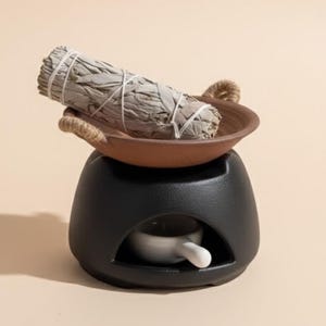 May include: A black ceramic essential oil diffuser with a small white dish and a brown ceramic bowl. A bundle of white sage is resting in the bowl. The diffuser has a rounded shape and a small opening.