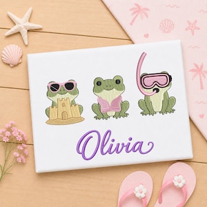 May include: A white fabric panel featuring an embroidered design of three green frogs. The first frog wears pink sunglasses and sits by a sandcastle. The second frog wears a pink life vest. The third frog wears a pink snorkel mask. The name Olivia is embroidered in purple script below.