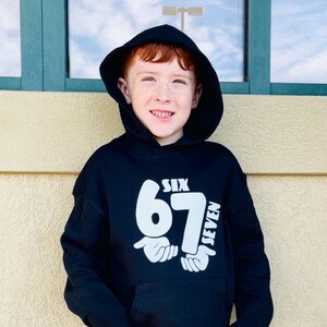 May include: A black hooded sweatshirt with the text "SIX 67 SEVEN" in white. The design includes the numbers 67 and the word "SIX" above the number 67, and the word "SEVEN" to the right of the number 67. The sweatshirt is worn by a person.