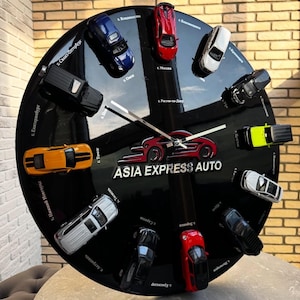 Könnte beinhalten: Eine schwarze Wanduhr mit Miniatur-Automodellen, die um das Zifferblatt angeordnet sind. Die Zeiger sind weiß, und der Text "ASIA EXPRESS AUTO" ist sichtbar. Die Uhr steht auf einem Tisch mit Backsteinmauer-Hintergrund.