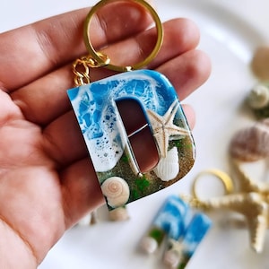 May include: A resin letter "D" keychain with a beach theme. The blue resin mimics ocean waves, with a starfish and seashells embedded. The keychain has a gold-colored ring.