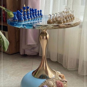 May include: A unique chess table with a round, resin top. One side features blue chess pieces, the other clear. The table has a gold-coloured stem and a light blue base adorned with seashells. The table is approximately 61 cm tall.