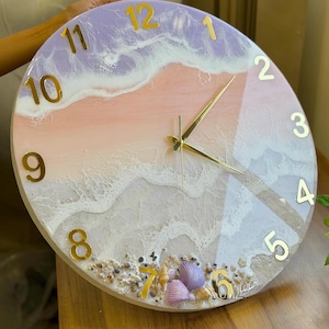 May include: A round wall clock featuring a resin art design with pastel lavender, peach, and white waves. The clock face includes gold-colored numbers from 1 to 12, gold hands, and a bottom section decorated with small seashells and pebbles.