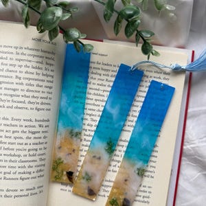 May include: Three handmade resin bookmarks with a beach-themed design. Each bookmark features a gradient of blue and white, resembling the ocean, with a sandy bottom and small decorative elements. They are displayed on an open book.