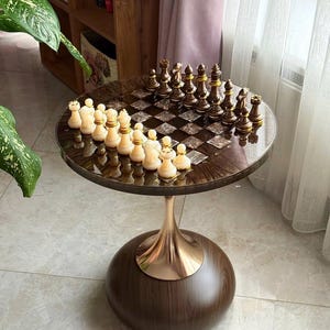 May include: A round chess table with a dark wood grain finish and a gold-colored base. The chess board is set with ivory and dark brown chess pieces. The table is on a tiled floor, near a window and a bookshelf.