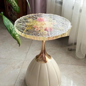 Puede incluir: Una mesa auxiliar decorativa con una tapa transparente y texturizada con flores incrustadas. La mesa tiene un tallo dorado y una base redondeada de color crema. La mesa mide aproximadamente 61 cm de altura.
