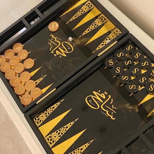May include: A black backgammon set with gold accents and Arabic calligraphy. The game board features gold triangles with decorative patterns. Tan and black game pieces with the letter "S" are visible, along with a die.