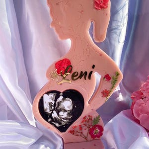 May include: A pink resin sculpture of a pregnant woman, decorated with dried flowers and the name "Leni". The sculpture includes a heart-shaped cutout showing a sonogram image. A unique keepsake.