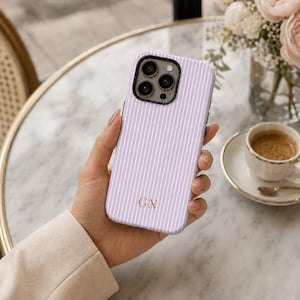 Peut inclure: Une coque de téléphone violet clair avec de fines rayures verticales blanches et les initiales GN de couleur dorée imprimées près du bas. La coque présente une découpe pour l'objectif de l'appareil photo et recouvre l'arrière du smartphone.