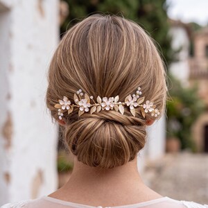 Può includere: Un pettine per capelli color oro con delicate foglie in metallo e piccoli accenti floreali bianchi con perle centrali. L'elemento decorativo presenta un motivo orizzontale a tralcio, progettato per essere inserito in un'acconciatura raccolta.