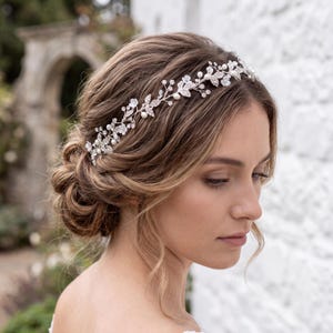 May include: A silver-toned hair vine featuring delicate floral motifs, small white pearls, and clear crystals arranged in a vine-like pattern. The flexible accessory is worn across the head, accentuating a styled updo with intricate metallic details.