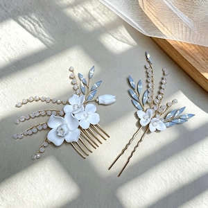 Puede incluir: Un peine para el cabello y una horquilla con detalles de flores blancas, hojas de cristal azul claro y pequeñas cuentas blancas sobre tallos de alambre dorado. El peine tiene varios dientes, mientras que la horquilla presenta un diseño de dos puntas.