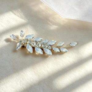 May include: A hair clip featuring a branch-like design with marquise-cut iridescent opal-toned crystals and a central row of clear round rhinestones set in a silver-toned metal frame, arranged in a leaf-like pattern extending from the center.
