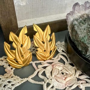 May include: A pair of gold-colored leaf-shaped earrings. The earrings have a textured surface and a clip-on style. The earrings are displayed on a gray surface with a white lace doily and a decorative amethyst crystal.
