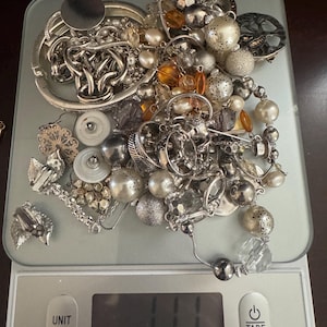 May include: A selection of silver-coloured jewellery, including necklaces, bracelets, earrings, and beads, is arranged on a digital kitchen scale. The scale reads 11.1, with the brand name "TAYLOR" visible.