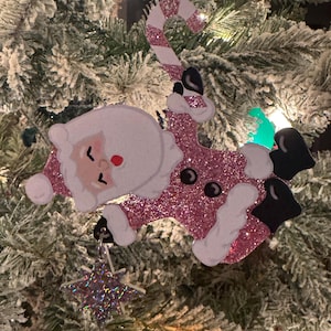 May include: A whimsical Christmas tree ornament featuring Santa Claus riding a reindeer, both adorned with pink glitter. The ornament includes a candy cane and a star-shaped charm, all hanging on a snowy Christmas tree.