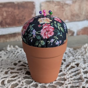 May include: A terracotta-colored pin cushion shaped like a flower pot. The cushion is covered in black fabric with a floral pattern of pink, red, and yellow flowers. Several colorful pins are inserted into the cushion.