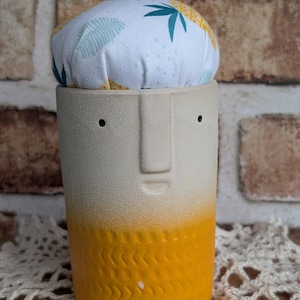 May include: A handmade ceramic pin cushion with a face design. The cylindrical base is cream-colored with a yellow textured bottom. The cushion is white with a tropical leaf pattern and holds several colorful pins.