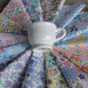 May include: A white teacup sits atop a fan of colorful floral fabric swatches. The fabrics display a variety of pastel floral patterns in shades of pink, blue, yellow, and purple. The teacup has a small handle and a simple design.