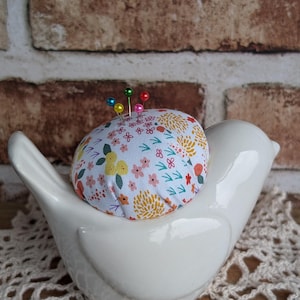 May include: A white ceramic bird-shaped pin cushion holder with a floral patterned pin cushion. The pin cushion has a white background with colorful flowers and green leaves. Several colorful pins are inserted into the cushion.