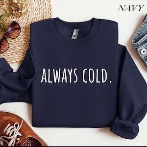 Può includere: Felpa blu navy con la scritta bianca "ALWAYS COLD". La felpa girocollo è realizzata in un materiale morbido. La parola "NAVY" si trova nell'angolo in alto a destra dell'immagine.